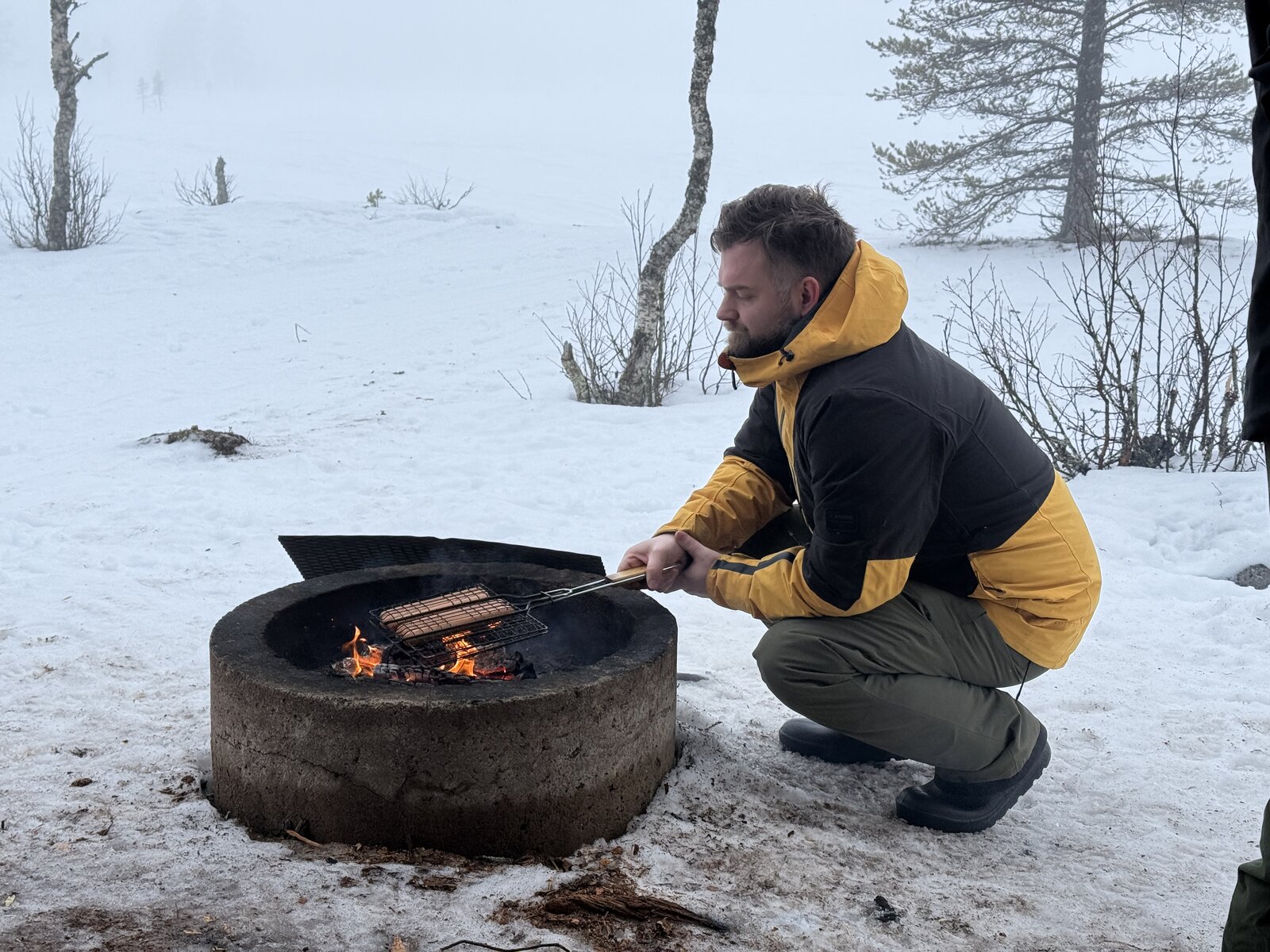 Martin grilling sausages over a fire pit in the snow